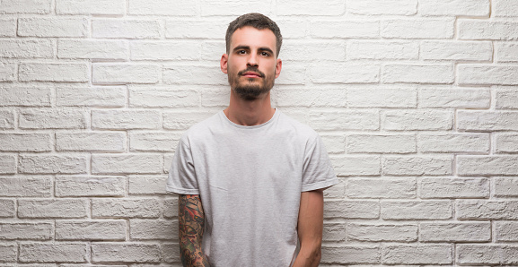 Young adult man standing over white brick wall with a confident expression on smart face thinking serious