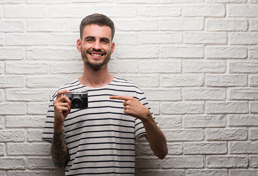Young man holding vintage camera standing over white brick wall very happy pointing with hand and finger