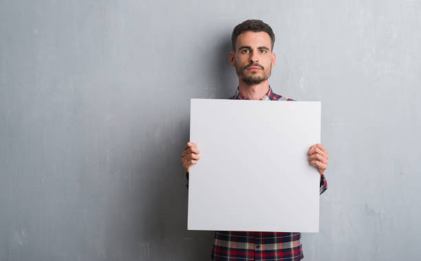 istock Young adult man over brick wall holding banner with a confident expression on smart face thinking serious 1042506388