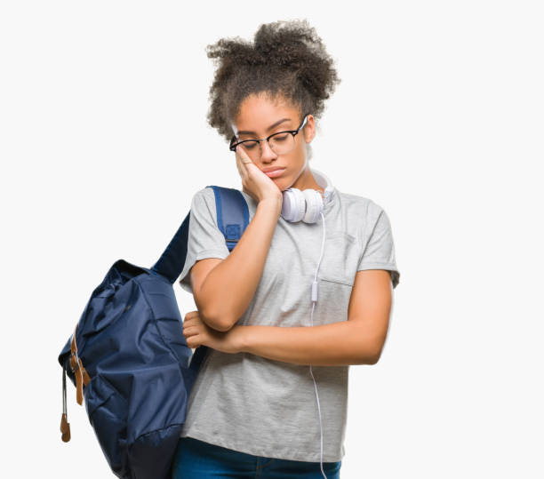 молодая афро-американская студентка в наушниках и рюкзаке на изолированном фоне мышления выглядит уставшей и скучной от проблем депрессии - arms crossed audio стоковые фото и изображения