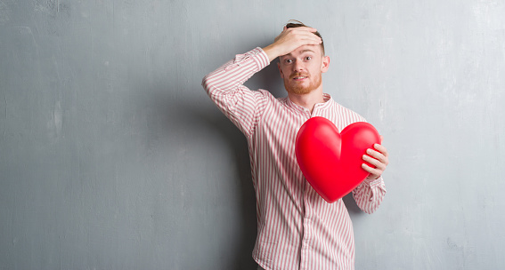 Young redhead man over grey grunge wall holding red heart stressed with hand on head, shocked with shame and surprise face, angry and frustrated. Fear and upset for mistake.