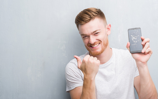 Young redhead man over grey grunge wall holding broken smartphone pointing and showing with thumb up to the side with happy face smiling