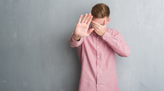 Young redhead man over grey grunge wall wearing pink shirt covering eyes with hands and doing stop gesture with sad and fear expression. Embarrassed and negative concept.
