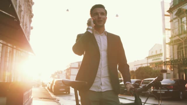 Handsome young stylish man talking on mobile phone outdoors with bicycle in city center during sunset