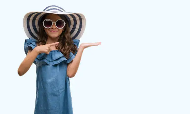 Photo of Brunette hispanic girl wearing sunglasses and summer hat very happy pointing with hand and finger