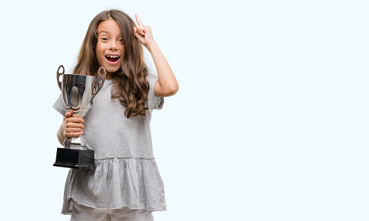 Brunette hispanic girl holding a trophy surprised with an idea or question pointing finger with happy face, number one