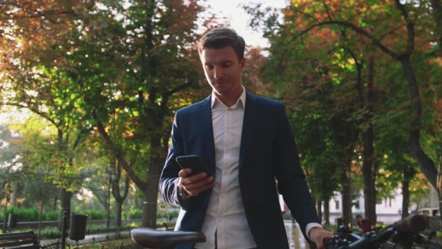 Handsome young stylish man using smartphone outdoors with bicycle in park in city center during sunset, slow motion, close up shot