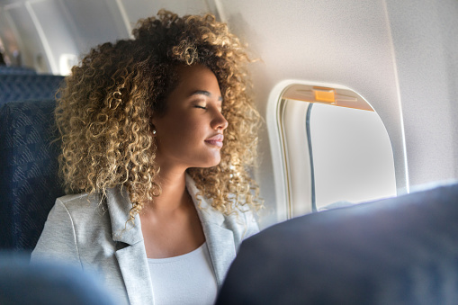 A young woman sits in the window seat of a commercial airliner and leans back with her eyes closed.