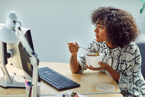 African businesswoman on lunch break in the office. Sitting by the desk, looking at monitor and eating her soup.