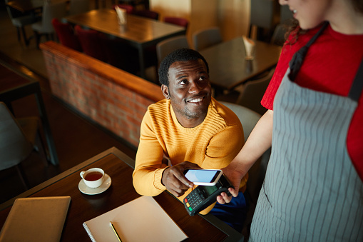Young smiling man sitting by table and looking at waitress while paying for his order through smartphone