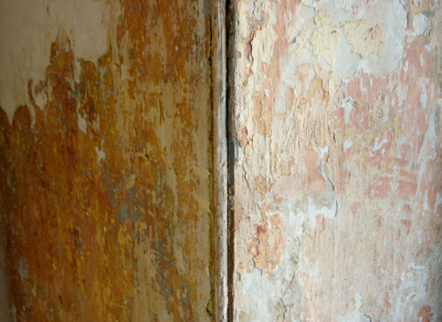 Stock photo showing close-up view of red peeling masonry paint layers on damp grey concrete wall with flaking plaster.