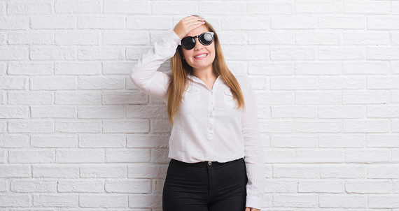 Young adult business woman standing over white brick wall wearing sunglasses stressed with hand on head, shocked with shame and surprise face, angry and frustrated. Fear and upset for mistake.