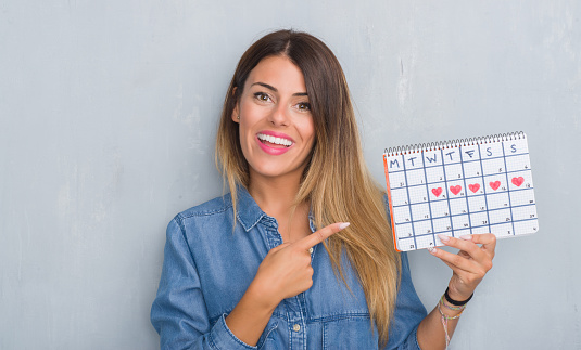 Young adult woman over grey grunge wall showing period calendar very happy pointing with hand and finger