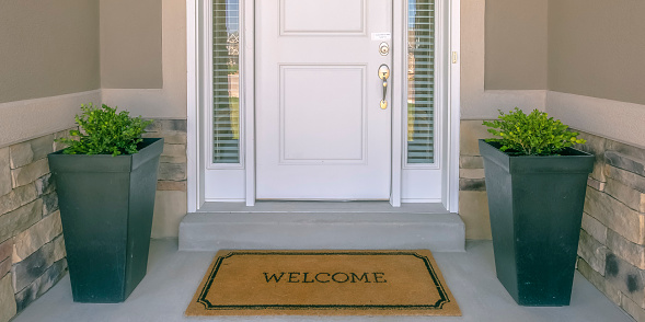 Front door with doormat plants and glass panel. White front door of a house with the word welcome printed on the brown doormat. Ornamental plants and glass panels are on each side of the door.