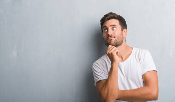 istock Handsome young man over grey grunge wall with hand on chin thinking about question, pensive expression. Smiling with thoughtful face. Doubt concept. 1042766184