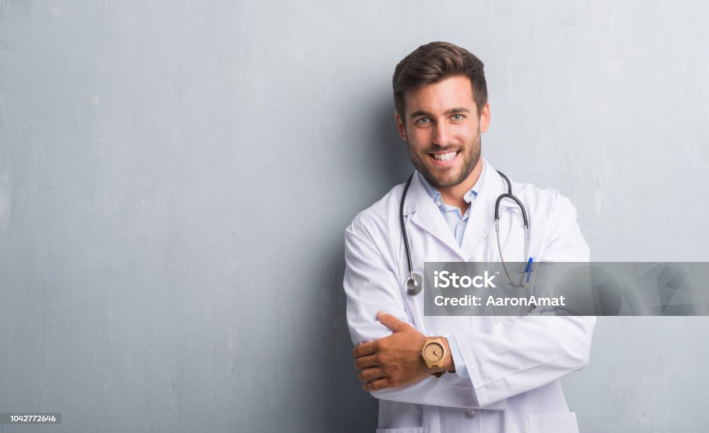 Handsome young doctor man over grey grunge wall happy face smiling with crossed arms looking at the camera. Positive person. Doctor Stock Photo