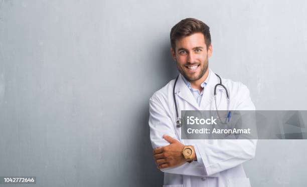 Handsome Young Doctor Man Over Grey Grunge Wall Happy Face Smiling With Crossed Arms Looking At The Camera Positive Person Stock Photo - Download Image Now