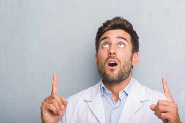 istock Handsome young professional man over grey grunge wall wearing white coat amazed and surprised looking up and pointing with fingers and raised arms. 1042772992