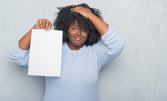 Young african american woman over grey grunge wall holding blank paper sheet stressed with hand on head, shocked with shame and surprise face, angry and frustrated. Fear and upset for mistake.