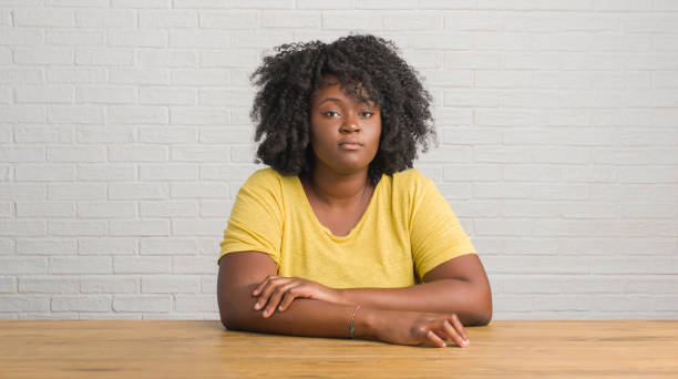 Young african american woman sitting on the table at home with serious expression on face. Simple and natural looking at the camera. Young african american woman sitting on the table at home with serious expression on face. Simple and natural looking at the camera. serious black teen stock pictures, royalty-free photos & images