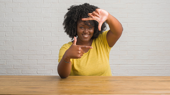 Young african american woman sitting on the table at home smiling making frame with hands and fingers with happy face. Creativity and photography concept.