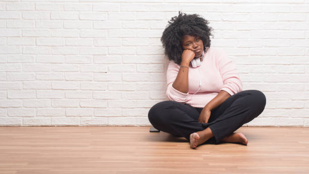 молодая афроамериканка, сидящая на полу в наушниках, думает, что выглядит уставшей и скучной от проблем с депрессией со скрещенными руками. - arms crossed audio стоковые фото и изображения