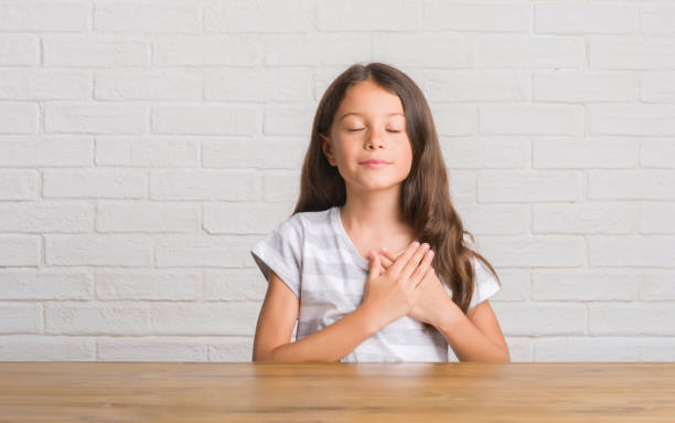 Young hispanic kid sitting on the table at home smiling with hands on chest with closed eyes and grateful gesture on face. Health concept. Young hispanic kid sitting on the table at home smiling with hands on chest with closed eyes and grateful gesture on face. Health concept. in pride we trust stock pictures, royalty-free photos & images