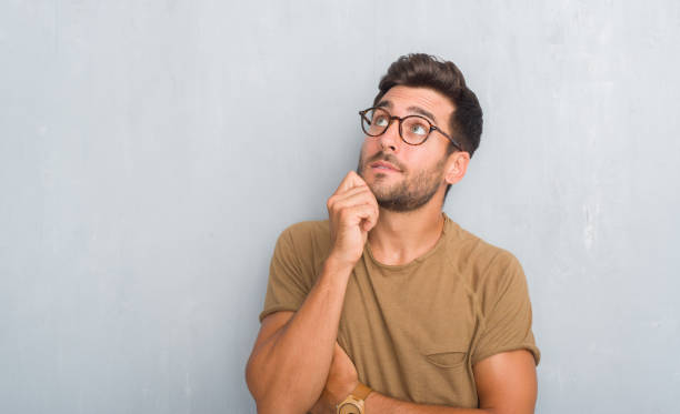 istock Handsome young man over grey grunge wall wearing glasses with hand on chin thinking about question, pensive expression. Smiling with thoughtful face. Doubt concept. 1042811652