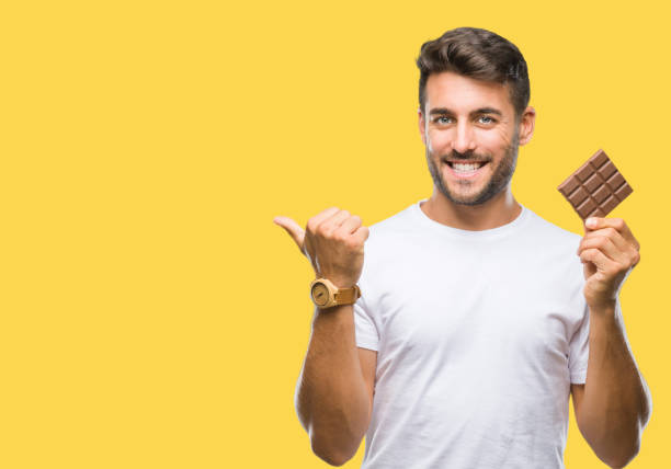 istock Young handsome man eating chocolate bar over isolated background pointing and showing with thumb up to the side with happy face smiling 1042816686