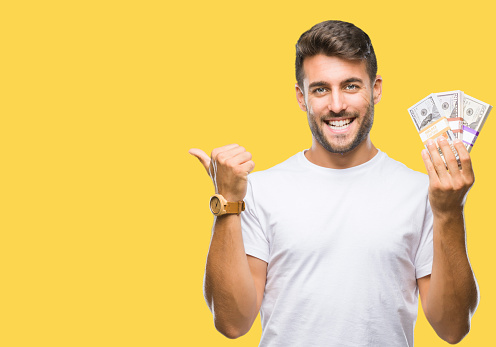 Young handsome man holding stack of dollars over isolated background pointing and showing with thumb up to the side with happy face smiling