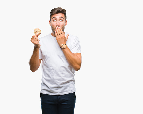 Young handsome man eating chocolate chips cookie over isolated background cover mouth with hand shocked with shame for mistake, expression of fear, scared in silence, secret concept