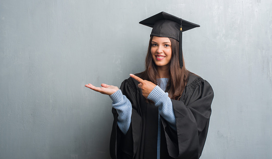 Young brunette woman over grunge grey wall wearing graduate uniform very happy pointing with hand and finger