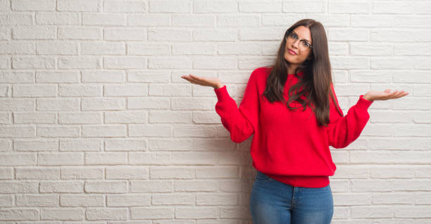istock Young brunette woman standing over white brick wall clueless and confused expression with arms and hands raised. Doubt concept. 1042824902