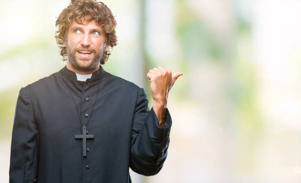 istock Handsome hispanic catholic priest man over isolated background smiling with happy face looking and pointing to the side with thumb up. 1042827376
