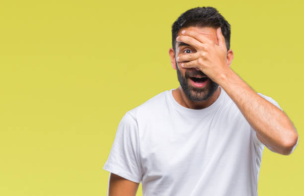 Adult hispanic man over isolated background peeking in shock covering face and eyes with hand, looking through fingers with embarrassed expression. Adult hispanic man over isolated background peeking in shock covering face and eyes with hand, looking through fingers with embarrassed expression. Hidden Meaning stock pictures, royalty-free photos & images