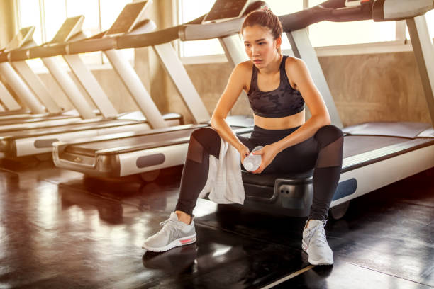 Beautiful Asian young woman tired taking a break from running or exercise sitting on treadmill machine drinking water and towel sweat in fitness gym healthy .girl in sportswear workout rest in morning stock photo