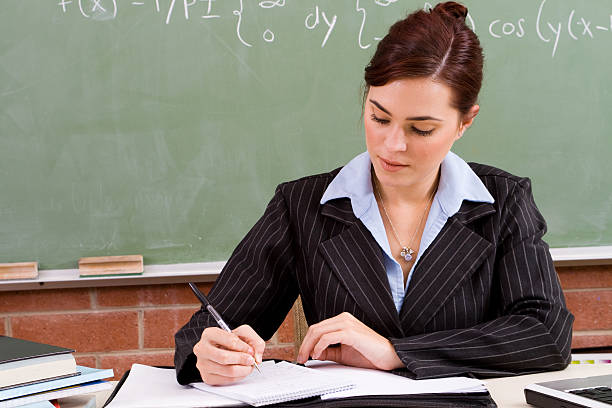 A mature African American high school teacher in class with students.  The teacher has short gray hair, wears black-framed glasses and a white suit sweater.  Her arms are folded in front of her holding a red folder.  Two boys and one girl are on the left, and one girl is on the right, all smiling.