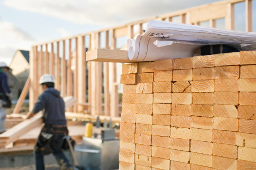 A closeup of stacks of 2x4 boards at a construction site, with a roll of blueprints sitting on top.  Two construction workers and a building frame can be seen in the background. Horizontal shot.