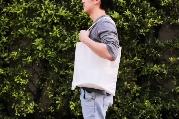jeune mâle hipster holding et transportant un sac vide de toile blanche sur fond de nature verte - espace de la copie. - tote bag photos et images de collection