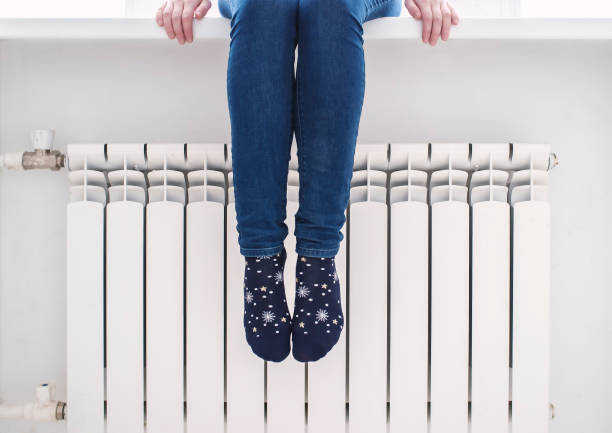 A girl sits on a window sill and warms the feet of the radiator, on a cold winter day. The girl sits on the windowsill and warms her feet in pretty socks at the radiator, on a cold winter day. warms stock pictures, royalty-free photos & images