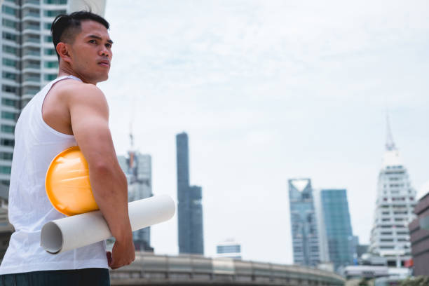 Asian engineer foreman worker with muscular holding yellow hardhat helmet in hand. Young handsome engineer man wear white vest shirt posing with construction site project on city background.