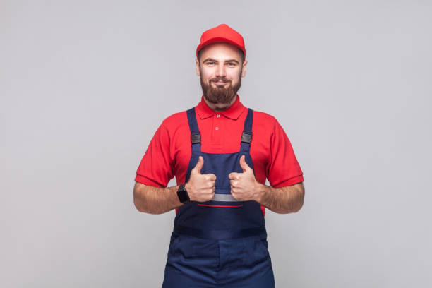 il lavoro è finito! ritratto di giovane riparatore allegro soddisfatto con barba in tuta blu, maglietta rossa e berretto, in piedi e mostrando tonf con il sorriso. - thumps up foto e immagini stock