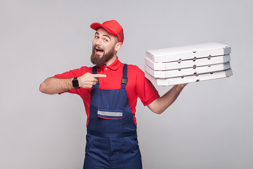 Young happy delivery man with beard in blue uniform and red t-shirt standing, holding and pointing finger to stack of cardboard pizza boxes on grey background. Indoor, studio shot, isolated.