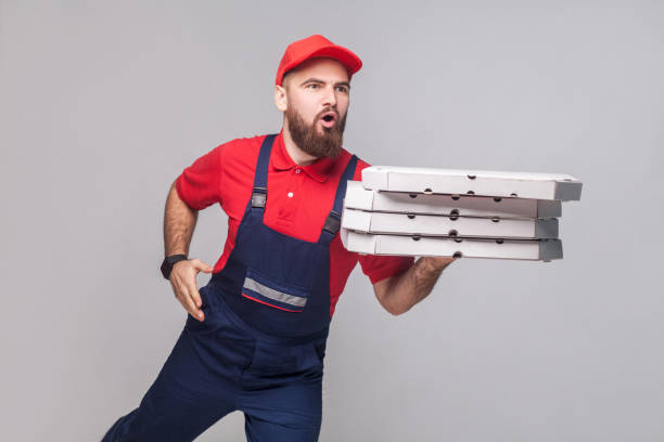 istock Hurry up! Young amazed delivery man with beard in blue uniform and red t-shirt holding stack pizza boxes and running to delivery on grey background. 1043036254