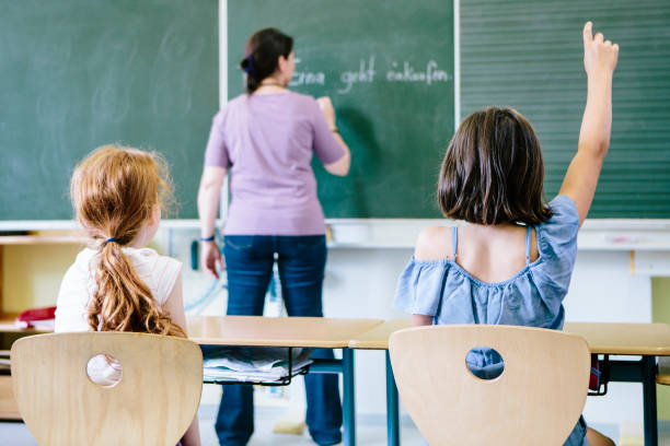 female teacher writes at the blackboard. rear view of two girls in front, on with raised arm. female teacher writes at the blackboard. rear view of two girls in front, on with raised arm. northern european stock pictures, royalty-free photos & images