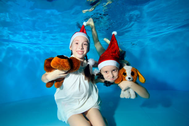 crianças no caps do papai noel são nadar e brincar debaixo d'água na piscina, segurando um cachorro de brinquedo - um símbolo de 2018, olhando para a câmera e a rir. retrato. visualização horizontal - underwater dog adult happiness - fotografias e filmes do acervo