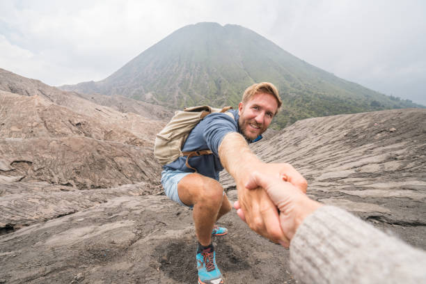 junger mann wandern, zieht hand eines teamkollegen zu erreichen. eine helfende hand an die spitze der vulkan krater erreichen. bromo vulkan region in indonesien, asien - bromo crater stock-fotos und bilder