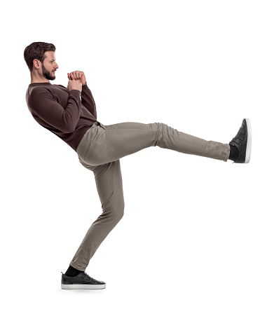 An isolated bearded man in casual clothes stands on white background in the middle of leg kick. Unexpected kick. Make opponent fall. Strong physique.