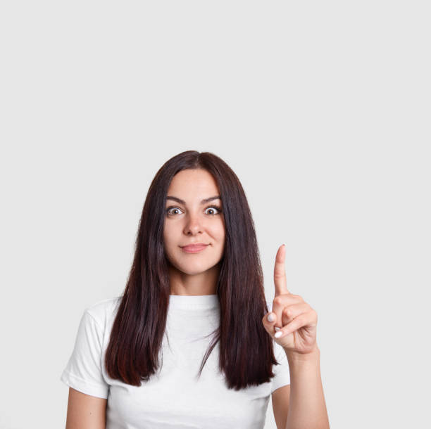 foto vertical de morena misteriosa mulher com longos cabelos escuros, aponta com o dedo indicador para cima, tem surpreendido a expressão, mostra algo incrível, sugere o uso de espaço em branco do seu anúncio - indexfinger - fotografias e filmes do acervo