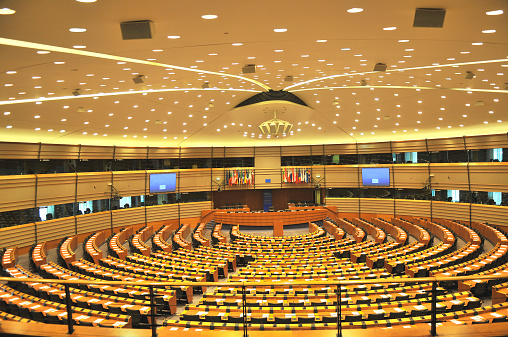 Empty big assembly room of European Parliament in Brussels, Belgium, Europe. Wooden chairs in semi-circle, papers on the banks, lots of boxes for translaiters, European flags and billboards in the background, many lights on the ceiling, fence in front. No people seen. Sandy brown color.
